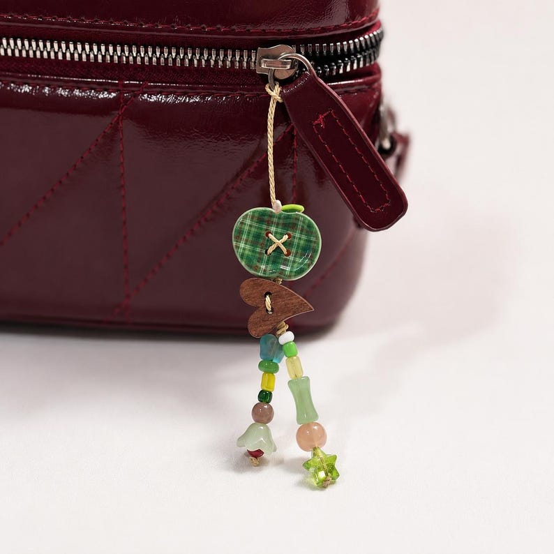 May include: A deep burgundy quilted bag with a zipper pull featuring a dangling charm. The charm consists of a green plaid apple-shaped button, a small wooden heart, and two strands of colorful glass beads in shades of yellow, green, and blue.