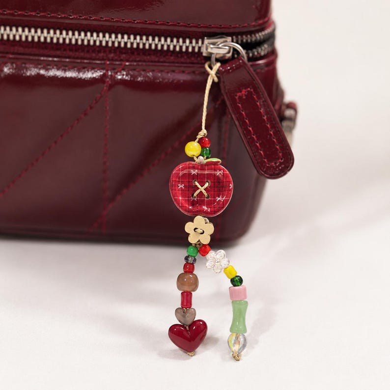 May include: A decorative bag charm attached to a dark red patent leather bag zipper. The charm features a red plaid apple-shaped button, a gold flower-shaped bead, and two dangling strands of colorful glass and plastic beads in heart and floral shapes.