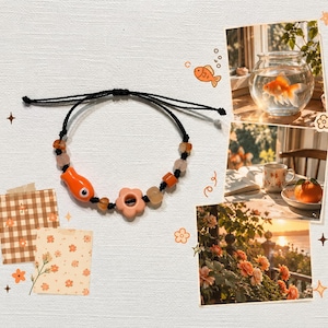 May include: A black cord bracelet featuring an orange fish-shaped bead, a light peach flower-shaped bead, and several smaller round orange and translucent beads. The bracelet is displayed against a background with decorative illustrations.