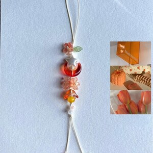 May include: A decorative beaded charm with a cream-coloured string. The charm features orange, yellow, and clear beads in the shape of flowers, a star, and a crescent moon. Likely for a phone or bag.
