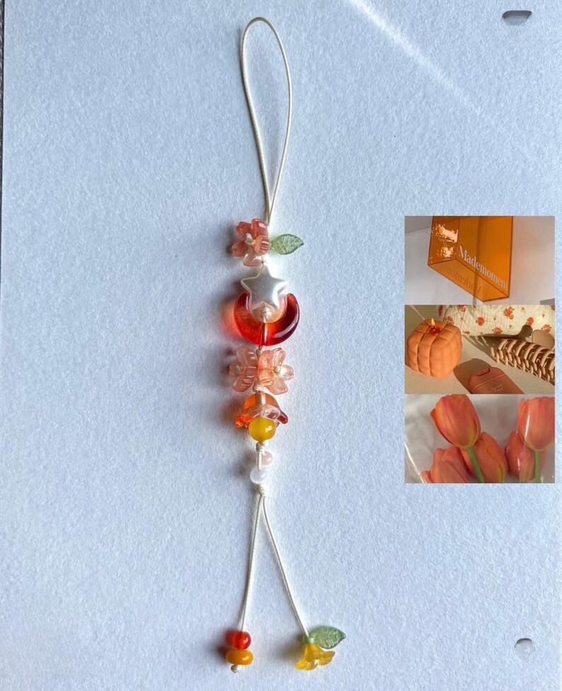 May include: A decorative beaded charm with a cream-coloured string. The charm features orange, yellow, and clear beads in the shape of flowers, a star, and a crescent moon. Likely for a phone or bag.