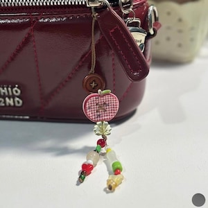 May include: A dark red quilted handbag with a silver zipper and a dangling charm featuring a brown button, a pink and white gingham apple-shaped pendant, a small clear leaf-shaped bead, and two strands of colorful glass and plastic beads.