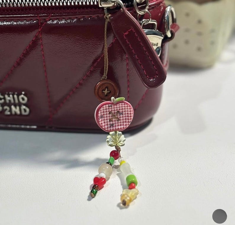 May include: A dark red quilted handbag with a silver zipper and a dangling charm featuring a brown button, a pink and white gingham apple-shaped pendant, a small clear leaf-shaped bead, and two strands of colorful glass and plastic beads.