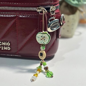 May include: A deep red quilted handbag with a silver zipper pull and a dangling charm featuring a green apple-shaped button, a beige button, and various small green, yellow, and clear beads arranged in a multi-strand design.