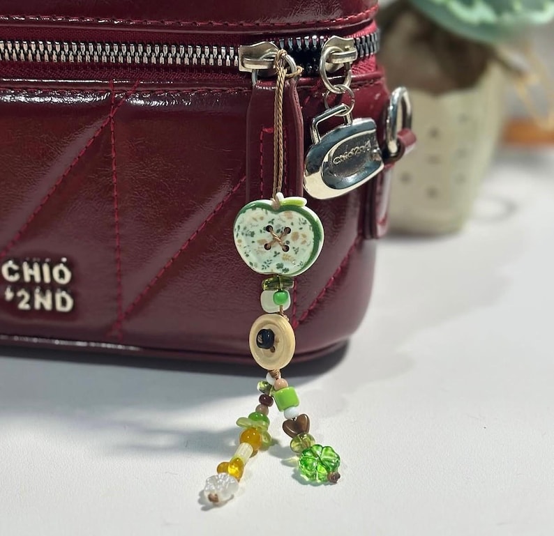 May include: A deep red quilted handbag with a silver zipper pull and a dangling charm featuring a green apple-shaped button, a beige button, and various small green, yellow, and clear beads arranged in a multi-strand design.