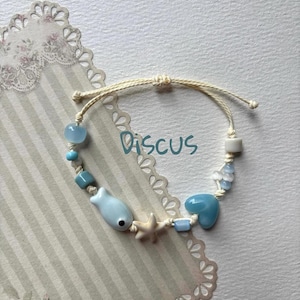 May include: A handmade bracelet featuring light blue beads, a fish charm, and a starfish charm. The bracelet is strung on a beige cord and includes the word "Discus". The bracelet is on a decorative background.