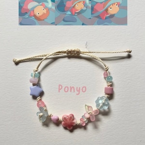 May include: A beaded bracelet on a light-coloured cord featuring the word Ponyo in pink text. The bracelet includes various beads in shapes of flowers, stars, and spheres in shades of pink, light blue, and lavender, arranged along the string.
