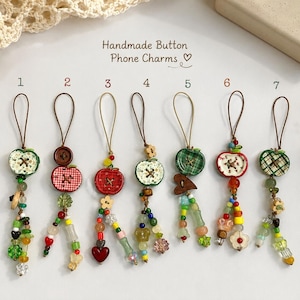 May include: Seven handmade phone charms labeled 1 through 7, featuring apple-shaped buttons, plaid and floral patterns, and colorful glass beads on brown cords. The text "Handmade Button Phone Charms" is printed above the items in a script font.