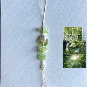 May include: A delicate green and white beaded phone charm. The charm features a loop, a star and moon pendant, flower-shaped beads, and small green and white beads. The charm is on a white string.
