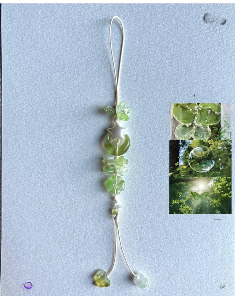 May include: A delicate green and white beaded phone charm. The charm features a loop, a star and moon pendant, flower-shaped beads, and small green and white beads. The charm is on a white string.