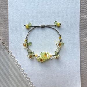 May include: A delicate bracelet featuring small yellow flower and leaf accents. The bracelet is strung on a grey cord and includes small yellow beads. The bracelet is displayed on a white card.