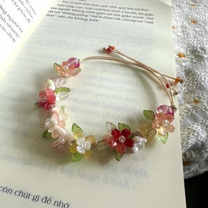 May include: A floral bracelet with pink, red, white, and yellow flower accents. The bracelet is made of beads and is resting on an open book. The book has text and the number 29 on the page.