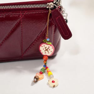 May include: A deep burgundy quilted patent leather pouch with a silver zipper closure and a decorative charm attached to the zipper pull. The charm features a white apple-shaped bead with red floral details and various colorful glass beads.