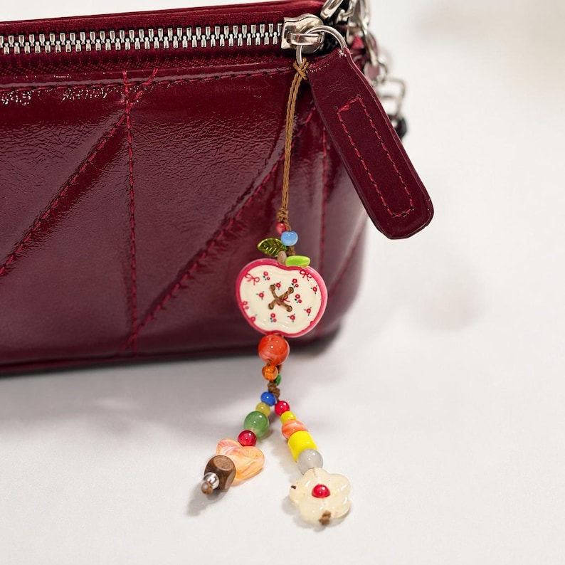 May include: A deep burgundy quilted patent leather pouch with a silver zipper closure and a decorative charm attached to the zipper pull. The charm features a white apple-shaped bead with red floral details and various colorful glass beads.
