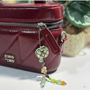 May include: A glossy dark red rectangular bag with a quilted geometric pattern and a silver zipper. The text CHIO 2ND is visible on the front. A decorative charm featuring a green plaid apple bead and various small green and yellow beads hangs from the zipper pull.