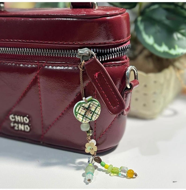 May include: A glossy dark red rectangular bag with a quilted geometric pattern and a silver zipper. The text CHIO 2ND is visible on the front. A decorative charm featuring a green plaid apple bead and various small green and yellow beads hangs from the zipper pull.