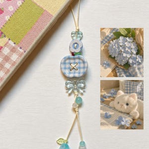 Puede incluir: Un colgante para teléfono con un botón en forma de manzana a cuadros vichy azul claro y blanco, una cuenta de lazo transparente, una cuenta de flor azul claro y un pequeño dije de tulipán azul en un cordón beige fino.