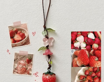 May include: A decorative hanging charm featuring a red strawberry pendant at the base, accented with a pink butterfly bead, green leaf beads, and a small pink bell-shaped bead, all strung on a thin brown cord with a loop at the top.