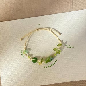 以下が含まれることがあります： ベージュのコードブレスレットで、花、葉、魚の形をした緑色のガラスビーズがあしらわれています。ブレスレットには小さな緑色のアクセントビーズが含まれており、デザインの周囲には緑色のインクでbướm、cá matcha、hoa lyという文字が書かれています。