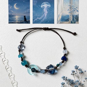 Puede incluir: Una pulsera de cordón marrón oscuro con un surtido de cuentas de vidrio azul y translúcido, que incluye una forma de media luna y un dije con forma de gato. La pulsera se muestra junto a tres impresiones artísticas cuadradas que representan un mar a la luz de la luna, una medusa y una vista de ventana con flores azules.