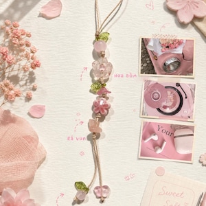 Könnte beinhalten: Ein dekorativer Hängeanhänger mit einer hellbraunen Kordel, auf die verschiedene rosa und klare Glasperlen in Blumen- und Fruchtform gefädelt sind, akzentuiert durch kleine grüne blattförmige Perlen, die in zwei separaten Strängen enden.