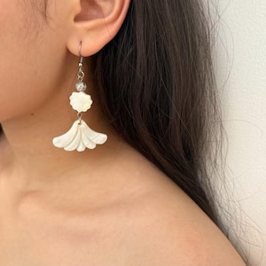 May include: White, shell-shaped earrings with a silver-colored hook. The earrings have a layered, fan-like design at the bottom and a small, round accent bead. The earrings are hanging from a person's ear.