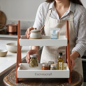 Puede incluir: Un estante de dos niveles de madera y blanco que contiene varios artículos de cocina. El estante superior tiene un tarro de cerámica con tapa, una bolsa de sal marina y una cuchara de madera. El estante inferior muestra botes de especias y una botella de aceite de oliva. La marca "Everoracraft" está impresa.