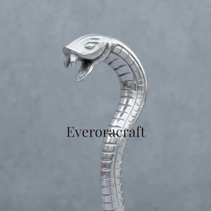May include: A polished silver-toned metal handle shaped like a cobra snake with its mouth open, showing visible fangs. The body of the snake features segmented scale detailing along its length. The text Everoracraft is printed in black.