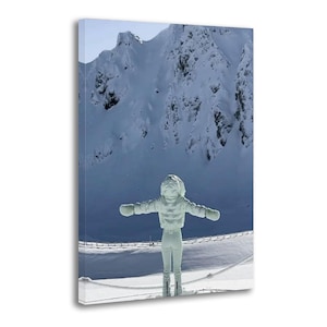 May include: A canvas print depicts a person in a white winter outfit with arms outstretched, standing in a snowy landscape. The background features a snow-covered mountain range under a clear sky. The person is wearing a hooded jacket, pants, and gloves.