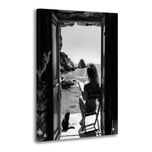 May include: A black and white wall art print depicting a view through an open doorway toward the sea. A person sits on a chair reading a book, while a cat sits on the floor nearby and another cat is perched on the side of the door frame.