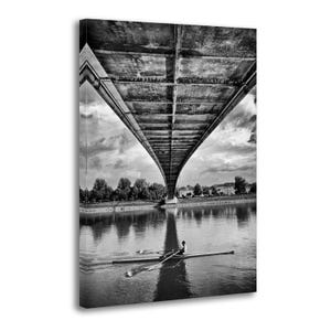 May include: Black and white photograph of a bridge arching over a river. A person in a rowing boat is in the water. The underside of the bridge is visible, with a cloudy sky in the background. Trees and buildings line the riverbank.