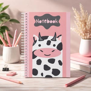 Puede incluir: Un cuaderno en espiral rosa con una ilustración de vaca de dibujos animados. La vaca tiene manchas negras y una nariz rosa. La palabra "Notebook" está en forma de nube gris oscuro. El cuaderno está sobre una superficie de madera clara.