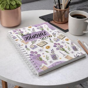 May include: A spiral-bound daily journal for 2026 with a white cover featuring a lavender and purple floral pattern. The journal includes illustrations of flowers, books, and lavender-infused drinks. The words "Daily Journal 2026" are printed on the cover.