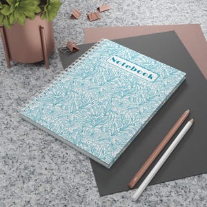 May include: A spiral-bound notebook with a teal floral pattern and the word "Notebook" on the cover. Two pencils, one white and one peach, lie beside the notebook. The notebook is on a gray surface with a plant and office supplies.