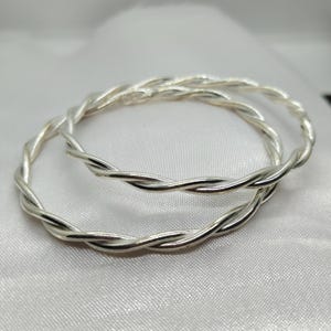 May include: Two silver-colored, twisted-rope design bangles. The bangles are circular and appear to be made of a shiny metal. The design consists of multiple strands of metal twisted together to form a rope-like pattern. The background is a white, silky fabric.