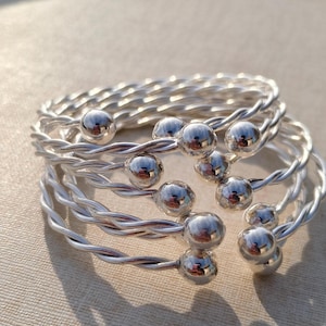 May include: A stack of silver-colored bracelets. Each bracelet features a twisted wire design and is adorned with a large, shiny, silver ball. The bracelets are stacked on top of each other, creating a layered effect. The overall aesthetic is modern and minimalist.