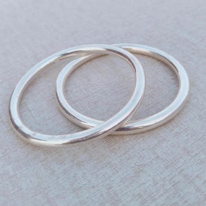 Può includere: Due braccialetti argentati, di forma circolare, sono interconnessi e appoggiati su uno sfondo neutro. I braccialetti hanno una finitura liscia e lucida, che riflette la luce. I gioielli sembrano essere in metallo.