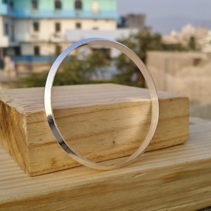 May include: A silver-colored bangle bracelet, circular in shape, resting on a wooden block. The bracelet has a smooth, polished surface and reflects the surrounding light. The background is blurred, with a building and greenery visible.