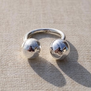 Puede incluir: Un anillo plateado con dos extremos esféricos pulidos. El anillo tiene un diseño curvo, con las esferas situadas en lados opuestos. El anillo está hecho de un metal brillante y reflectante, y proyecta una sombra sobre la superficie.
