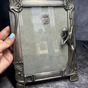 Könnte beinhalten: Silberfarbener Jugendstil-Bilderrahmen mit rechteckiger Form. Der Rahmen hat verzierte Eckdesigns und eine Skulptur einer Frau auf der rechten Seite. Der Rahmen ist etwa 15 cm hoch.