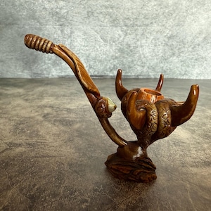 May include: A handcrafted wooden tobacco pipe with a distinctive design. The pipe has a curved stem with a textured grip, a bowl supported by a detailed, carved base, and a decorative element. The colour is a warm brown with lighter accents.