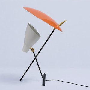 May include: A mid-century modern table lamp with a white cone-shaped shade and an orange, curved shade. The lamp has a black metal frame with brass accents and a black power cord. The lamp is approximately 20 inches tall.