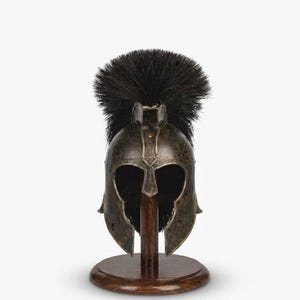 May include: A bronze-toned metal helmet featuring a wide, dark, textured crest made of black bristles. The helmet has a T-shaped face opening and is mounted on a circular, polished dark brown wooden stand with a central vertical support post.