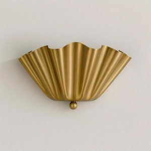 May include: A gold-colored wall sconce with a scalloped, fan-like design. The sconce has a rounded base and a textured surface. The fixture is made of metal and is mounted on a white wall.