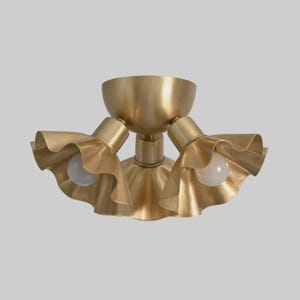 Può includere: Un lampadario da soffitto color ottone con tre paralumi a balze. Ogni paralume contiene una lampadina. Il lampadario ha una base centrale a forma di ciotola e bracci regolabili. Il design è di ispirazione vintage.