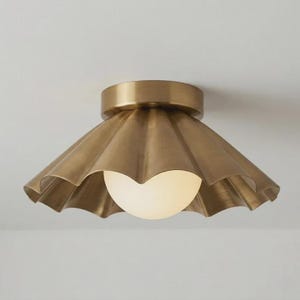 Puede incluir: Una lámpara de techo de montaje empotrado con una pantalla de latón festoneada y un acabado metálico cepillado. Una bombilla esférica de cristal blanco está centrada dentro de la pantalla, proporcionando una fuente de luz suave y difusa.