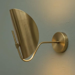 May include: A brass wall sconce featuring a curved, fan-shaped shade and a circular base. The sconce has a minimalist design and is mounted on a light-coloured wall. The fixture is made of metal and has a warm, golden finish.