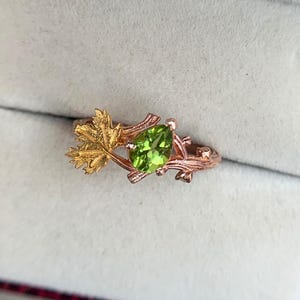 May include: A rose gold ring with a pear-shaped, light green gemstone. The ring features a leaf and branch design, with a gold-coloured leaf accenting the gemstone. The ring is set in a white jewellery box.