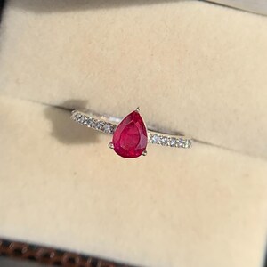 May include: A silver ring featuring a pear-shaped, deep red gemstone. The ring's band is embellished with small, clear gemstones. The ring is displayed in a brown jewellery box with a beige interior.