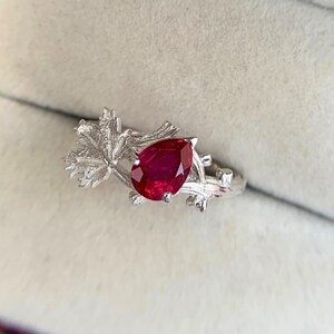 May include: A silver ring featuring a pear-shaped red gemstone set within a band designed to resemble textured tree branches and leaves. The metal has a matte, brushed finish, and the vibrant red stone is positioned as the central focal point.